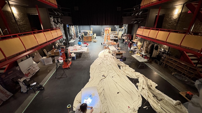 Vue d'ensemble de la salle de spectacle de la cité francophone, remplie de décorations, costumes, bâches. Des hommes et femmes travaillent sur des tables.