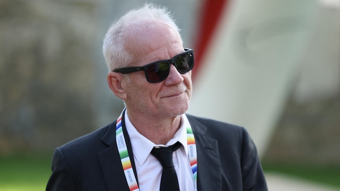 Un homme aux cheveux blancs portant des lunettes de soleil sourit. 