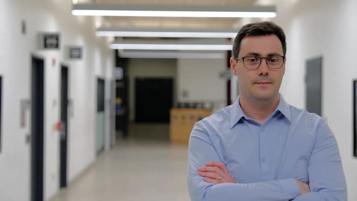Un homme portant une chemise et des lunettes croise les bras dans les couloirs d'une université.