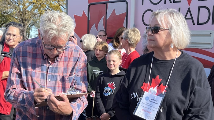 Un homme aux cheveux gris est en train d'enregistrer des informations pour une collecte de signatures, à l'extérieur. À sa gauche, une femme aux cheveux blond clair se tient à côté de lui. Plusieurs autres personnes se tiennent derrière eux.