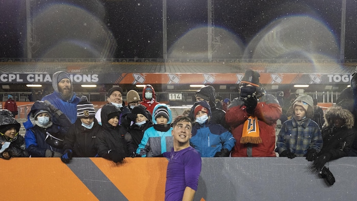 Un joueur vêtu d'un maillot mauve, après un match, est debout sur un tabouret pour s'approcher d'un groupe de spectateurs habillé chaudement pour le temps froid.