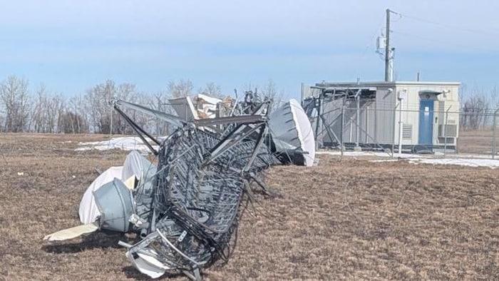 Une antenne de télécommunication effondrée au sol dans un champ partiellement enneigé, avec en arrière-plan un bâtiment technique entouré d'une clôture.