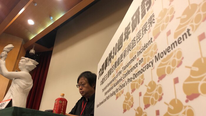 Un homme est attablé à la tribune d'une conférence à Taïwan. Et derrière lui se trouve une réplique de la « Déesse de la Démocratie ».