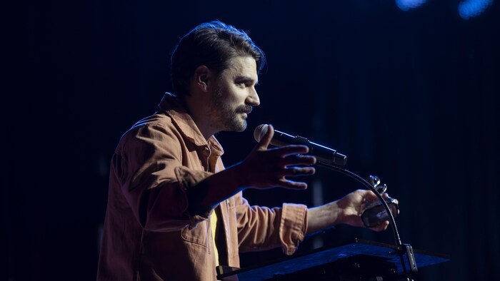 Un homme de profil, sur une scène, les bras ouverts, devant un micro.