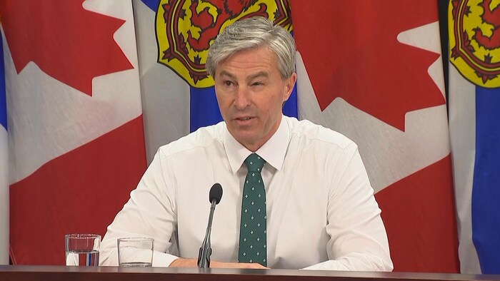 Tim Houston en chemise blanche et cravate verte à pois blancs, assis à une table brune, devant des drapeaux du Canada et de la Nouvelle-Écosse.