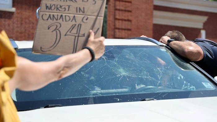 Des craques sur le pare-brise d'un véhicule routier blanc. Un homme qui semble être un policier en uniforme et dont on ne voit pas le visage regarde à l'intérieur du véhicule par une portière ouverte.