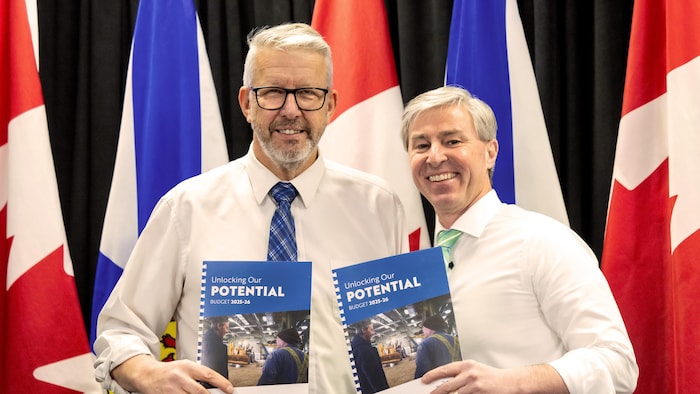 Vêtus de chemises blanches, John Lohr et Tim Houston posent côte à côte en souriant et tenant des brochures reliées du budget 2025-2026.