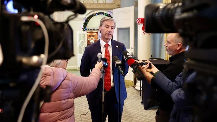 Le premier ministre portant une cravate rouge très festive et trop longue. Il a les deux bras dans le dos, est debout sous une couronne de Noël et est questionné par des journalistes.