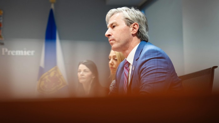 Le profil gauche de Tim Houston, assis à une table, l'air sérieux, à côté de deux femmes.