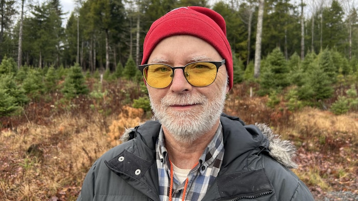 Un homme coiffé d'une tuque rouge et portant des lunettes aux verres jaunes est debout dehors devant des conifères.