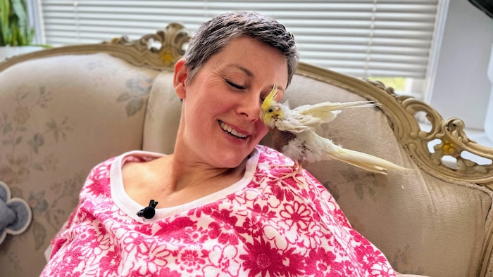 Une femme est assise sur un canapé avec un oiseau affectueux sur l’épaule.
