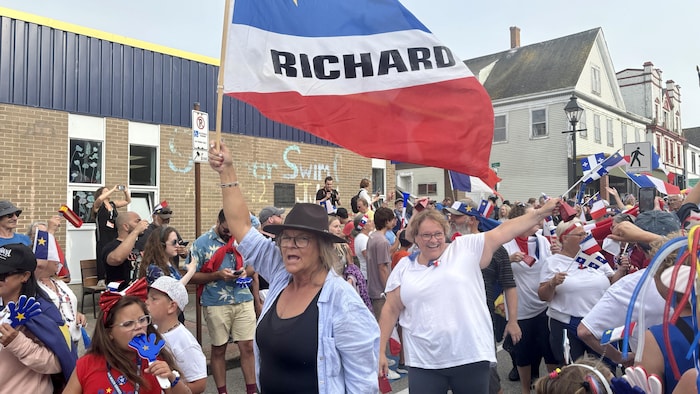 Dans un défilé, une femme coiffée d'un chapeau brun vocifère en agitant un gros drapeau aux couleurs de l'Acadie et décoré du nom « Richard » en lettres noires. Près d'elle, une fillette mord à belles dents dans un jouet en plastique bleu représentant une main.