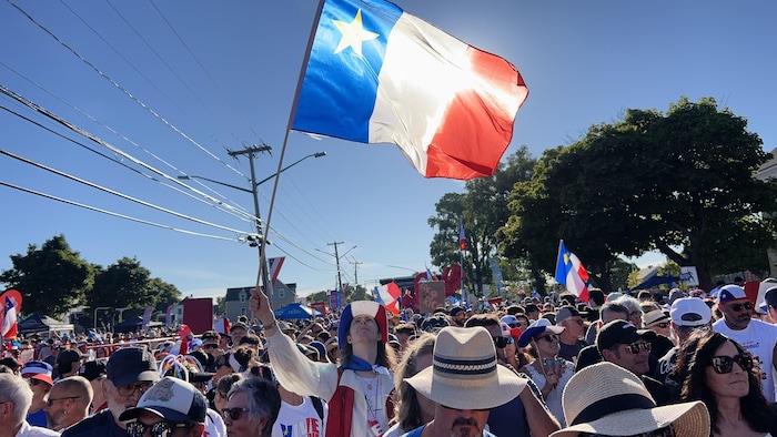 Une foule de gens habillés de bleu, blanc et rouge font du bruit sur la rue principale de Caraquet. Un homme lève un drapeau acadien dans le ciel bleu. 