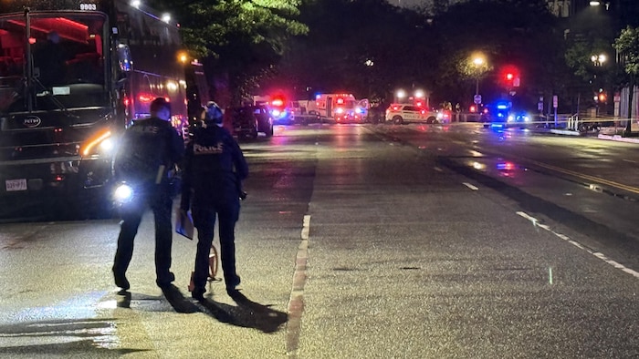 Des policiers marchent dans la rue, où se trouvent de nombreux véhicules d'urgence.
