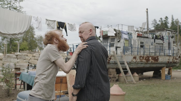Un homme agrippe un autre homme par l'épaule devant un bateau délabré servant d'habitation.