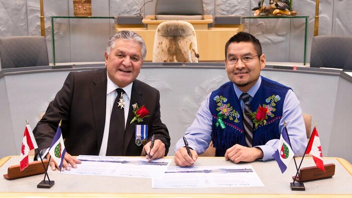 Deux homme signent des documents sur une table sur laquelle sont posés les drapeaux du Canada et des Territoires du Nord-Ouest.