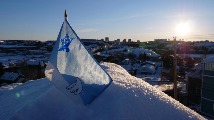 Un petit drapeau bleu et blanc arborant les symboles d'un ours polaire et d'un logo représentant une moitié de fleur de lys et une moitié de flocon est planté sur une butte de neige. En arrière-plan, la ville de Yellowknife.