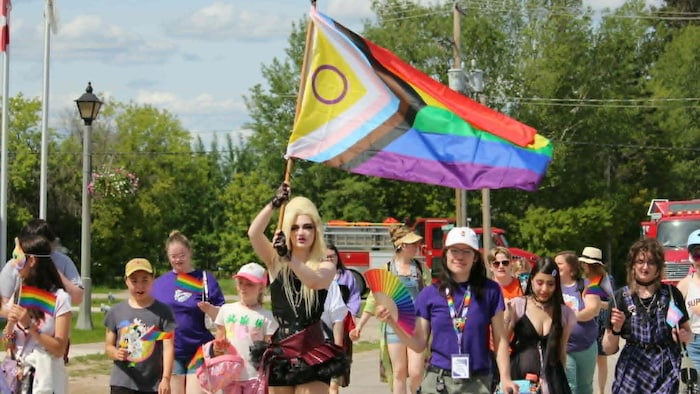 Une personne en tête d'une parade tient un grand drapeau arc-en-ciel.