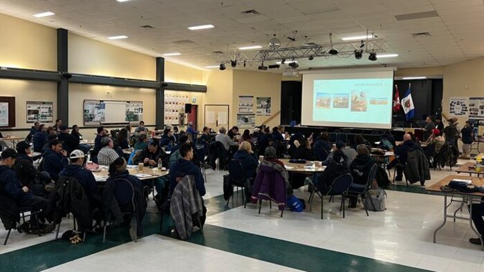 Une salle où des personnes sont réunies autour de plusieurs tables rondes face à un vidéo projecteur.