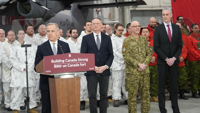 Mark Carney pendant sa conférence de presse devant des militaires et entouré de responsables politiques.