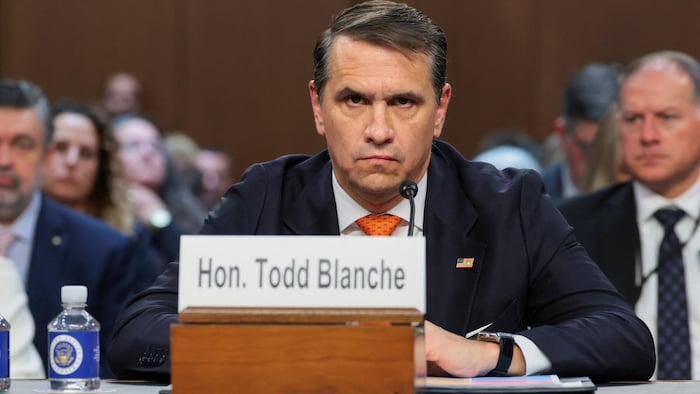 Todd Blanche, le regard sérieux, est assis derrière un microphone et une plaque nominative à son nom.