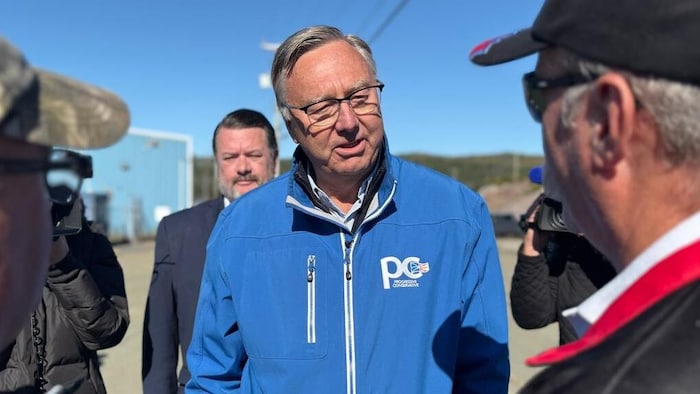Vêtu d'un blouson bleu, Tony Wakeham s'entretient avec des citoyens, à l'extérieur sous un ciel clair.