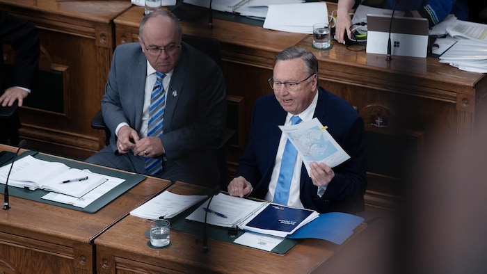 Deux hommes assis à la Chambre d'assemblée, l'un brandissant un document.