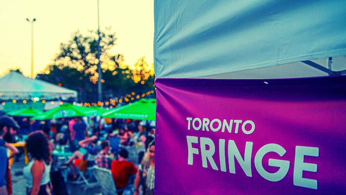 Au premier plan, un panneau qui nomme le Toronto Fringe. Au second plan, une foule assise sur des chaises sous des parasols.