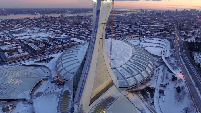 On voit le stade olympique du côté nord, de haut, en contre-plongée, l'hiver, au soleil couchant. Au loin, on aperçoit le fleuve Saint-Laurent et le centre-ville de Montréal.