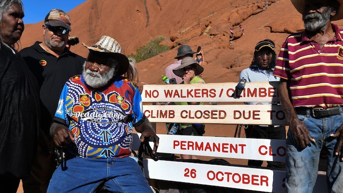 Des Aborigènes sont assis devant une montagne rouge et des inscriptions qui indiquent que la randonnée est interdite.