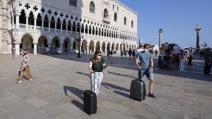 Des touristes portant des masques et des lunettes de soleil tirent leur valise sur le parvis de la place Saint-Marc.