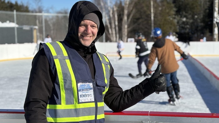 Un homme en veste réfléchissante sourit, le pouce en l'air, devant une patinoire où des gens jouent au hockey.