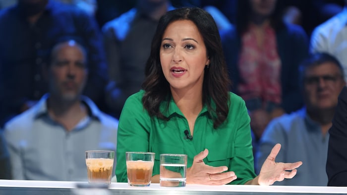 Ruba Ghazal en chemise verte s'exprime avec des gestes, assise à une table de studio avec trois verres, devant un public.