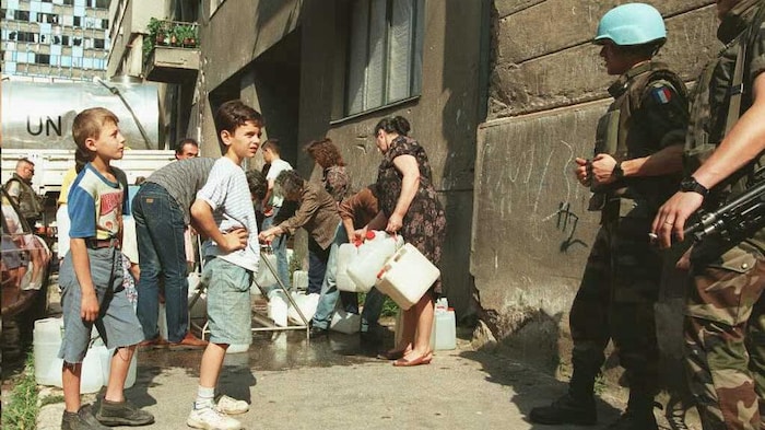 Des gens remplissent des bidons d'eau sous le regard de deux militaires.