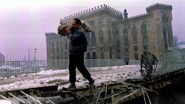 Un homme porte un sac de bois de chauffage sur un pont détruit, près de la bibliothèque nationale de Sarajevo, incendiée en 1994.