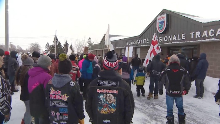 Des manifestants brandissent des pancartes et des drapeaux devant un édifice municipal. Deux amateurs de heavy metal portant des blousons d'Iron Maiden observent la scène.