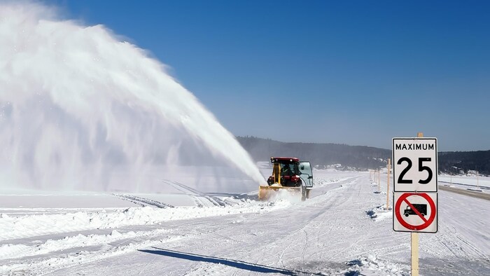 Tracteur qui souffle la neige sur un pont de glace qui relie deux rives d'un lac. 