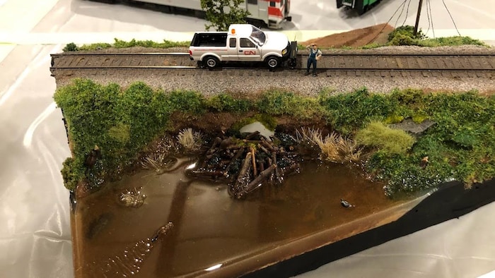 Un modèle miniature représente un rail de chemin de fer avec un véhicule arreté. Un homme miniature observe en se grattant la tête une sortie d'eau bouchée par les bois d'un castor.