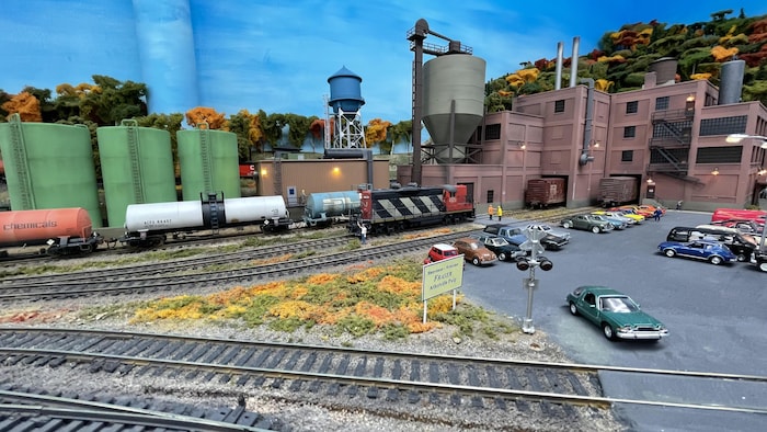 un décor miniature représente des arbres,un bâtiments industriel, des rails de chemin de fer et un stationnement avec des voitures.