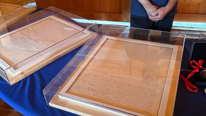 Des feuilles présentant une écriture et des signatures, encadrées dans une boîte transparente afin d'assurer la protection des papiers.
