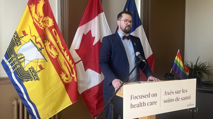 Un homme parle dans un micro, des drapeaux du Nouveau-Brunswick, du Canada et de l'Acadie derrière lui.