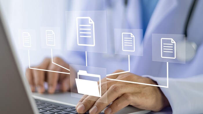 Deux mains d'homme manipulent le clavier d'un ordinateur portable. Sur la photo, des icônes dessinées représentent des fichiers et des documents pour illustrer le concept d'un transfert de fichiers par ordinateur.