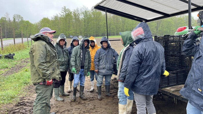 Bruno Paquette se tient debout dans la boue, avec les travailleurs vêtus de manteaux et de bottes de pluie.