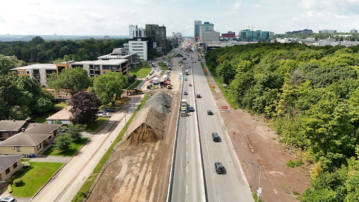 Vue par drone de l'artère bordée d'un côté par un quartier résidentiel et de l'autre par une petite forêt. Au loin, de grandes tours commerciales. Des tas de terre et de gravier recouvrent tout un côté du boulevard et la circulation est déviée à certains endroits. 