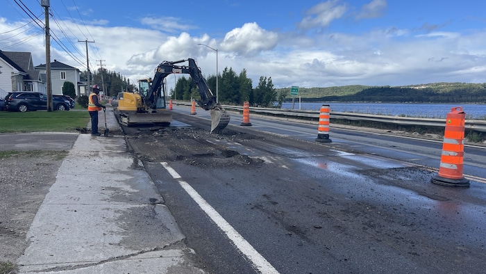 Une pelle mécanique est opérée sur une route asphaltée bordée de cônes.