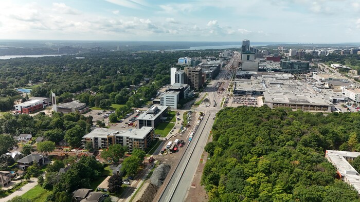 Vue par drone de l'artère bordée d'un côté par un quartier résidentiel et de l'autre par une petite forêt. Au loin, de grandes tours commerciales. Des tas de terre et de gravier recouvrent tout un côté du boulevard et la circulation est déviée à certains endroits. 