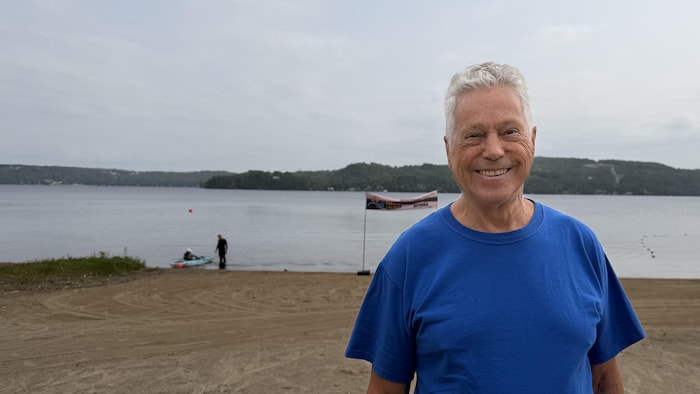Un homme âgé aux cheveux gris et au t-shirt bleu sourit au premier plan. En arrière-plan, une plage de sable borde un vaste lac entouré de collines boisées, avec une banderole et une personne près de l'eau sous un ciel nuageux.