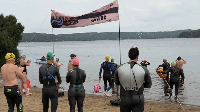 Plusieurs nageurs vêtus de combinaisons et de bonnets de bain sont rassemblés sur une plage au bord d'un lac. Une banderole « 10 km du Lac Otis » est tendue au-dessus du bord de l'eau sous un ciel nuageux.