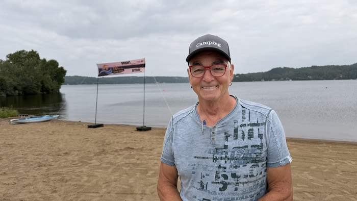 Un homme souriant portant une casquette « Camping » et des lunettes rouges se tient sur une plage de sable. En arrière-plan, un lac, une bannière « départ / arrivée » et un kayak sont visibles sous un ciel nuageux.