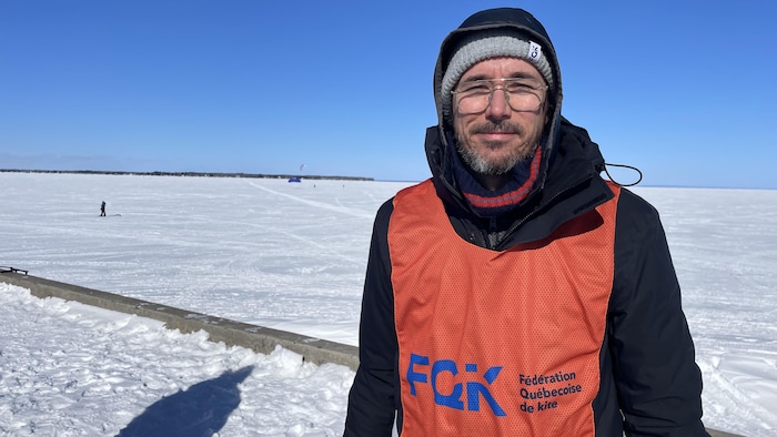 Un homme portant un dossard orange de la « Fédération Québécoise de kite » pose devant une vaste étendue enneigée sous un ciel bleu clair. On aperçoit au loin une silhouette et une voile de snowkite sur la surface glacée.